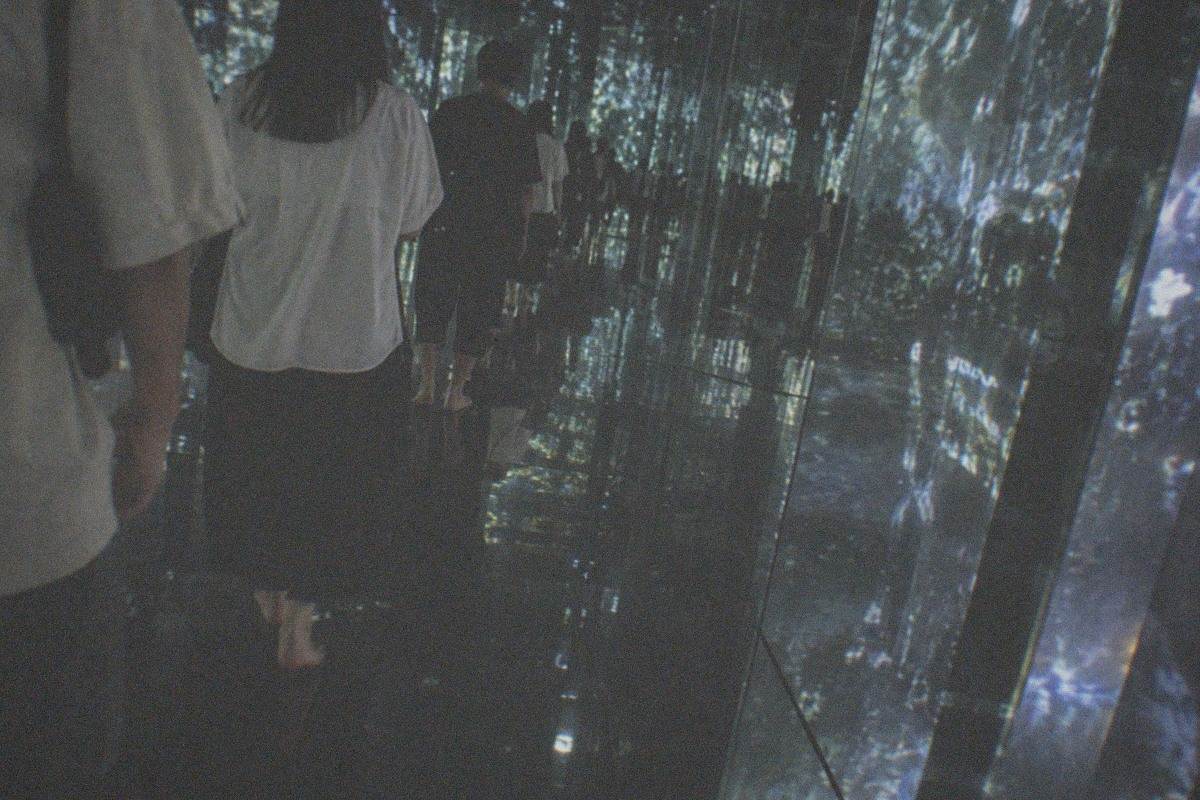 A dark gallery space at teamLab Planets with visitors wading through a shallow reflective water room lit by projected light