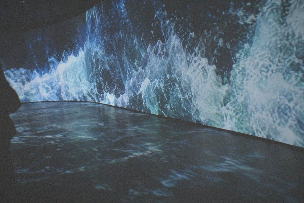 A dark museum gallery washed in shifting digital light projections across walls and floor